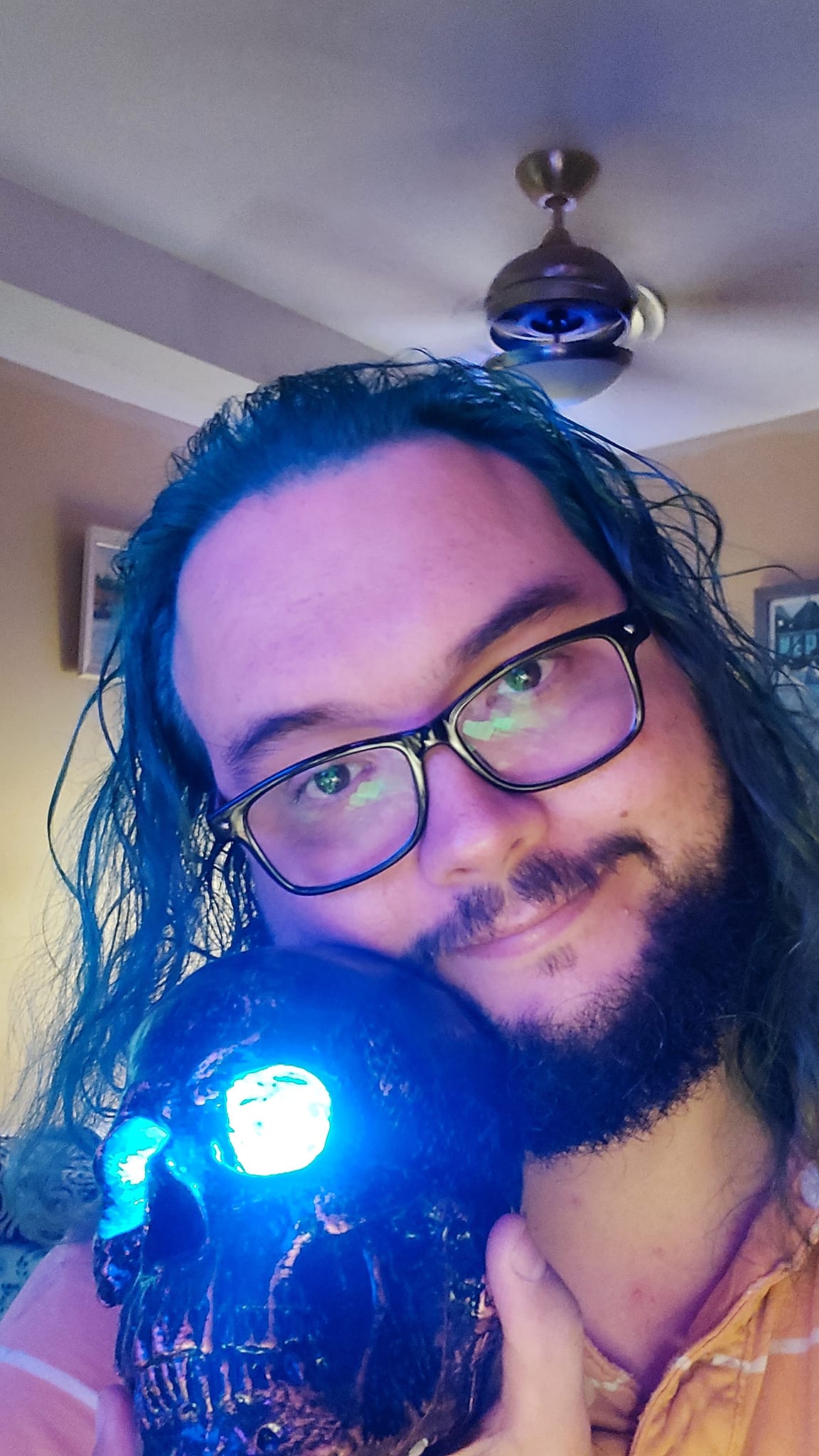 A man with long green hair sits in a computer chair holding a plastic skull